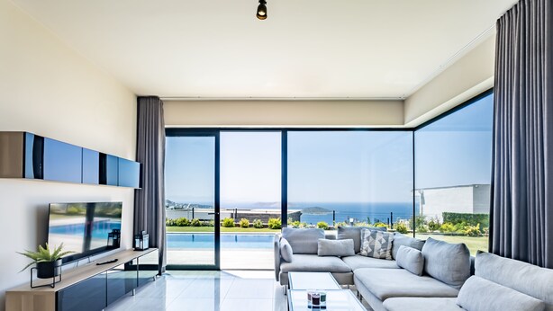 A modern living room with a large flat-screen TV, a sofa set, a coffee table, and a view of a swimming pool and ocean.
