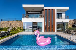Una casa moderna de dos pisos con piscina, un flotador con forma de flamenco rosa y mobiliario exterior.