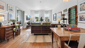 A spacious living area with a wooden coffee table, a sofa, and a dining table. There are framed pictures on the walls, a large window with curtains, and a hanging lamp. - Downtown Apartment! Large, Cozy, Family friendly private oasis in city center! (Petoskey)