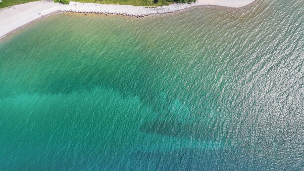 An aerial view of a beach with clear turquoise water, a sandy shore, and some greenery. - Downtown Apartment! Large, Cozy, Family friendly private oasis in city center! (Petoskey)
