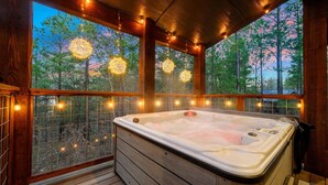 A hot tub with a wooden deck and glass walls, surrounded by string lights and a view of trees. - Cozy, Game Deck, Outdoor Swing, 2 King Suites, Save 7% in Taxes! (Broken Bow)