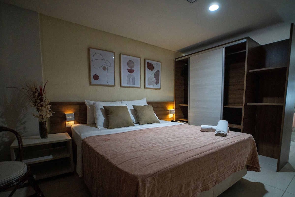 Una habitación de hotel con cama, mesitas de noche, una silla, escritorio, armario y cuadros en la pared.