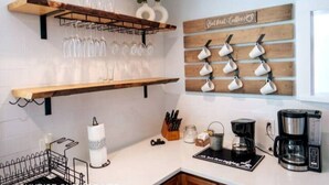 Une cuisine avec des étagères en bois où sont rangés des verres à vin et des tasses à café, une machine à café et un bloc à couteaux.