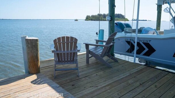 Une terrasse en bois avec deux fauteuils Adirondack, un bateau nommé «KEL WEST», et une vue sur un lac paisible avec une île au loin.