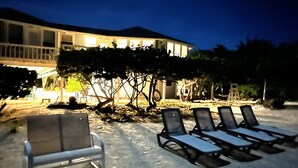 Un área de descanso junto a la playa con tumbonas y una silla blanca, un árbol y un edificio con ventanas iluminadas al fondo.