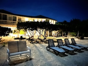 A beachside lounge area with sun loungers and a white chair, a tree, and a building with illuminated windows in the background. - Sonscape's Cozy 1-Bdr 1-Bath Private Guest Suite Opens Right Onto the Beach! (Cayman Brac)