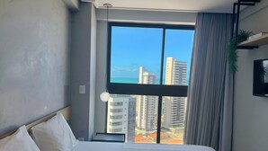 A hotel room with a large window, a bed with white linens, a mounted TV, and a plant on the windowsill. - Miramar One Way Tambaú (João Pessoa)