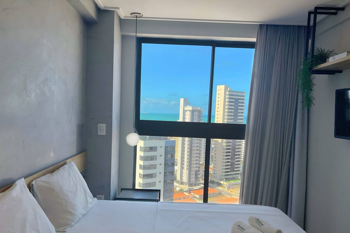 Een hotelkamer met een groot raam, een bed met witte lakens, een wandtelevisie en een plant op de vensterbank.