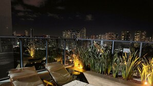 A rooftop terrace with lounge chairs and a table, overlooking a city skyline at night. - Miramar One Way Tambaú (João Pessoa)