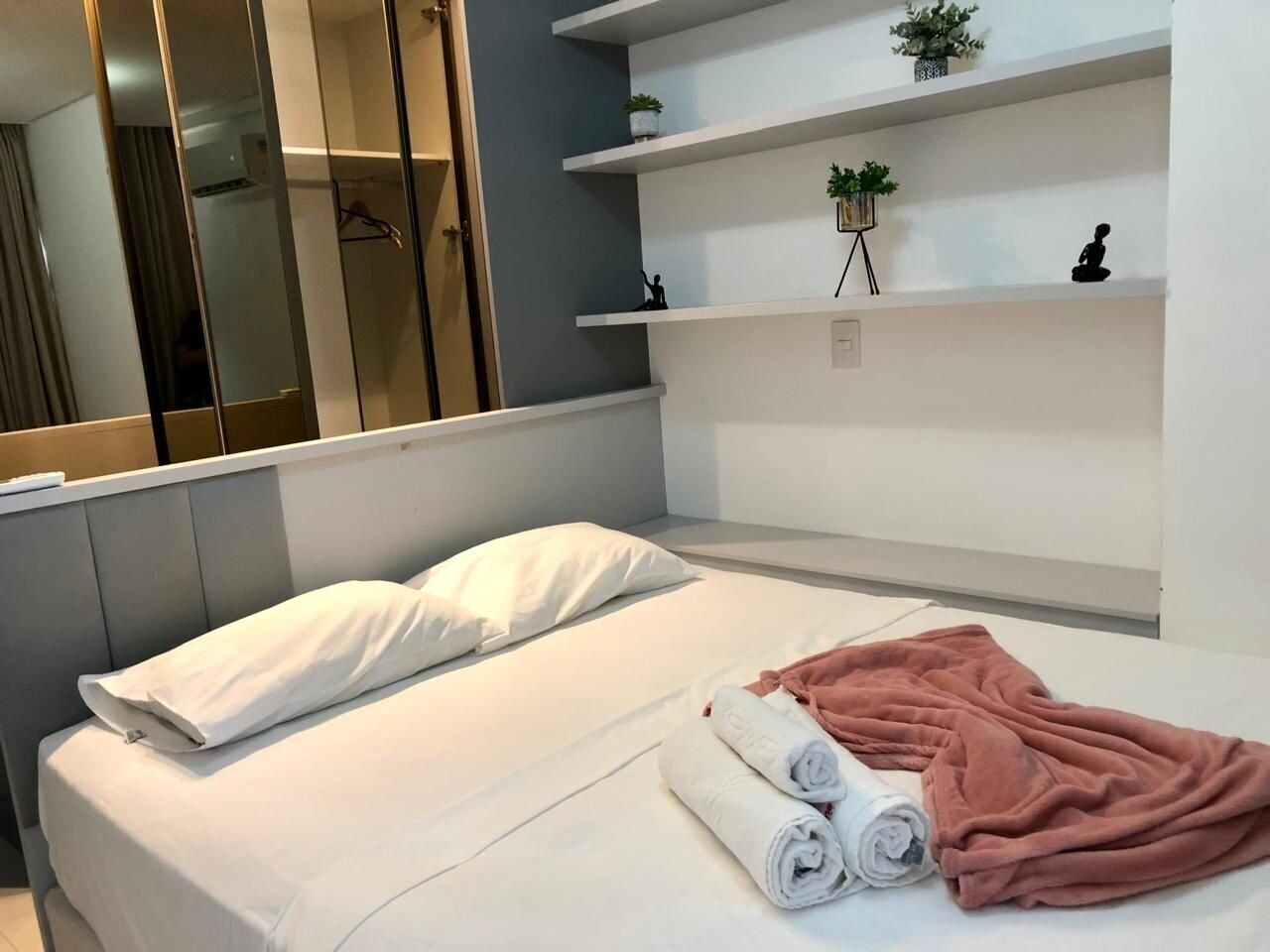 Cama bem arrumada com roupa de cama branca e rosa, uma pequena planta em uma prateleira e um espelho refletindo parte do quarto.