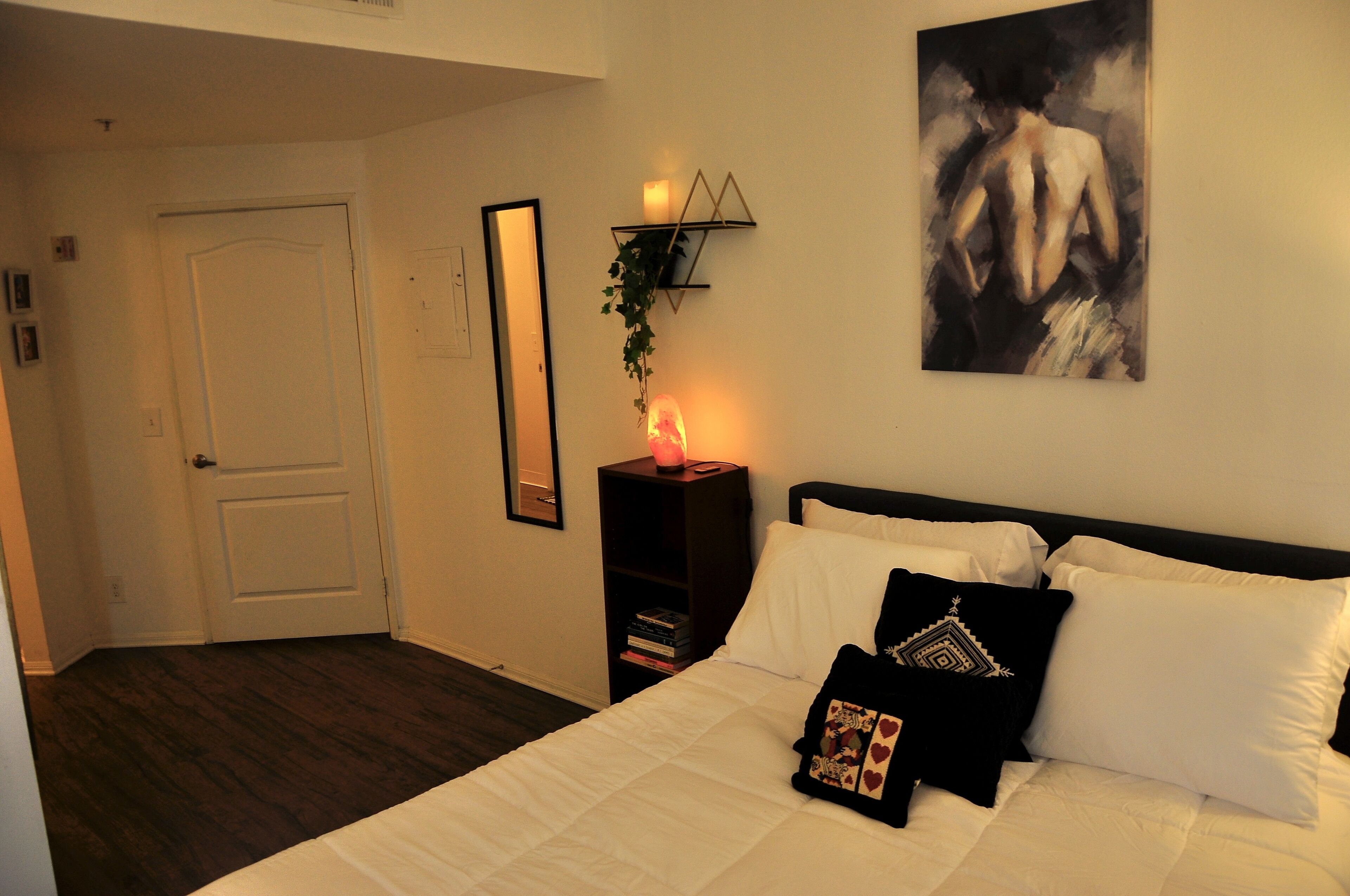 Ein Schlafzimmer mit einem Bett, weißer Bettwäsche, einem Nachttisch mit Kerze, einem gerahmten Bild an der Wand und einer Tür.