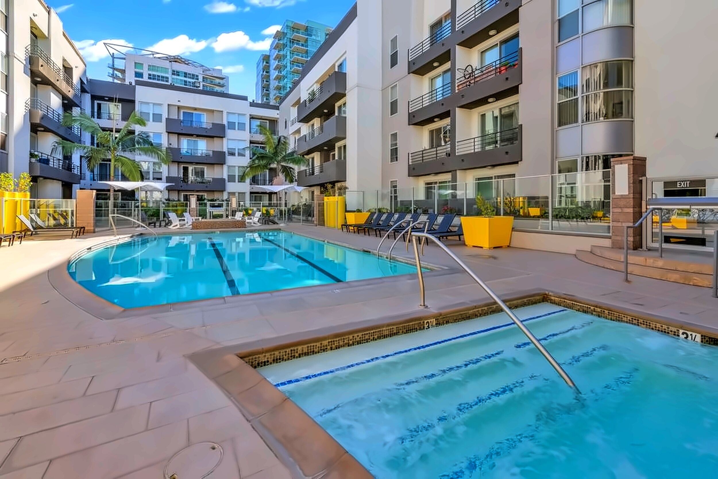 Une piscine dotée de plusieurs couloirs, entourée de chaises longues et de palmiers, avec un immeuble d’appartements moderne en arrière-plan.
