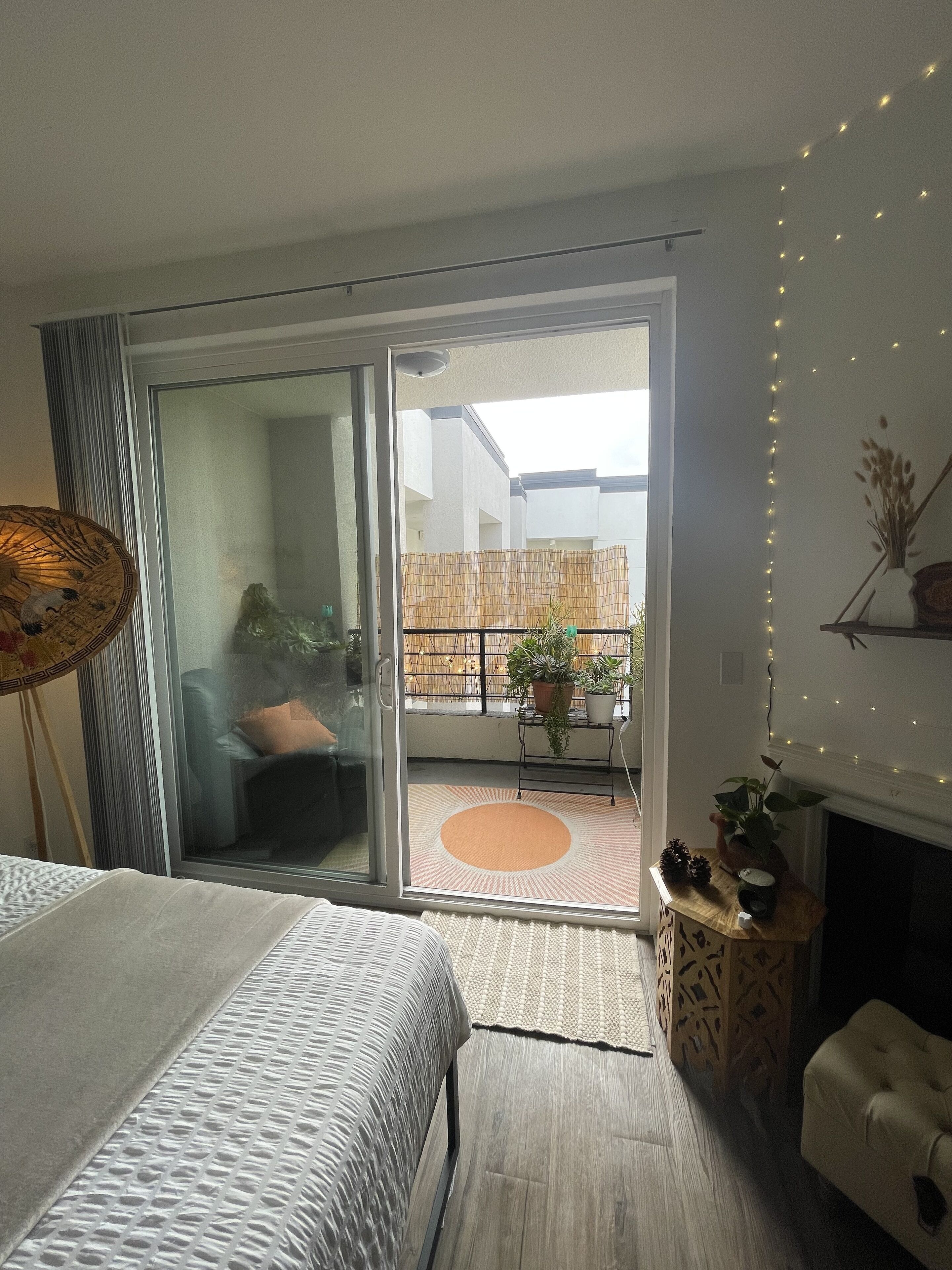 Un dormitorio con cama, puerta corrediza de vidrio que da a un balcón con plantas y vistas de un edificio.