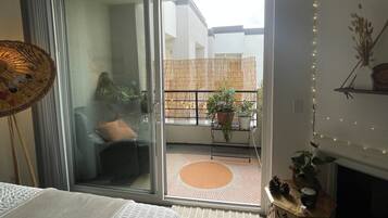 Un dormitorio con cama, puerta corrediza de vidrio que da a un balcón con plantas y vistas de un edificio.