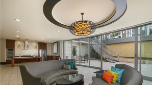 Una sala moderna con un sofá gris curvo, cojines coloridos, una mesa de centro de vidrio y una zona de cocina con horno y refrigerador integrados.