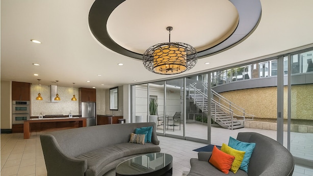 Una sala moderna con un sofá gris curvo, cojines coloridos, una mesa de centro de vidrio y una zona de cocina con horno y refrigerador integrados.