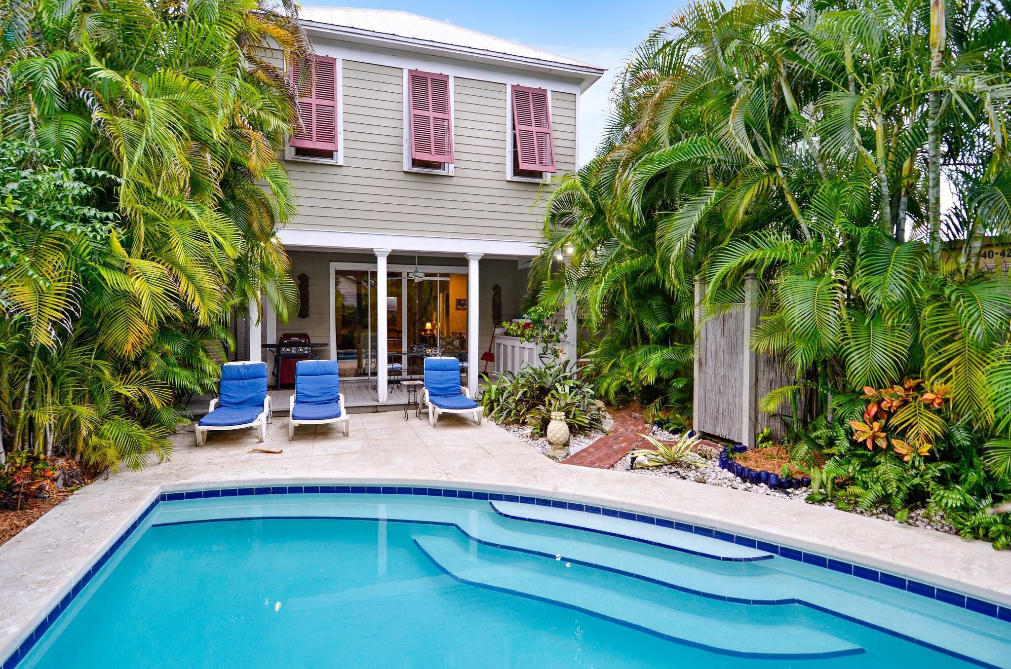 A house with a swimming pool, two lounge chairs, and tropical plants.