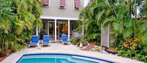A house with a swimming pool, two lounge chairs, and tropical plants.