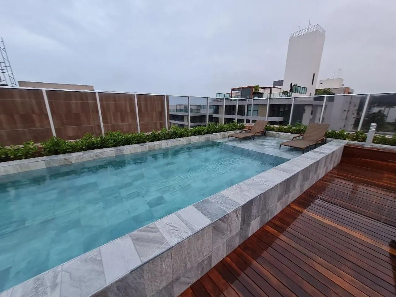 A rooftop swimming pool with a wooden deck, surrounded by a low wall and greenery.