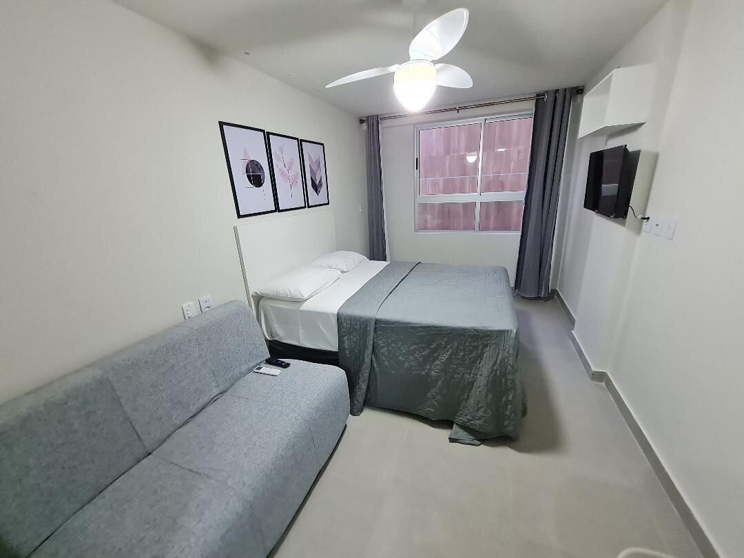 A small, well-lit room with a bed, a sofa, a ceiling fan, a window with curtains, and a wall-mounted TV.