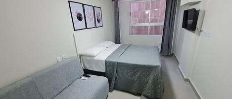 A small, well-lit room with a bed, a sofa, a ceiling fan, a window with curtains, and a wall-mounted TV.