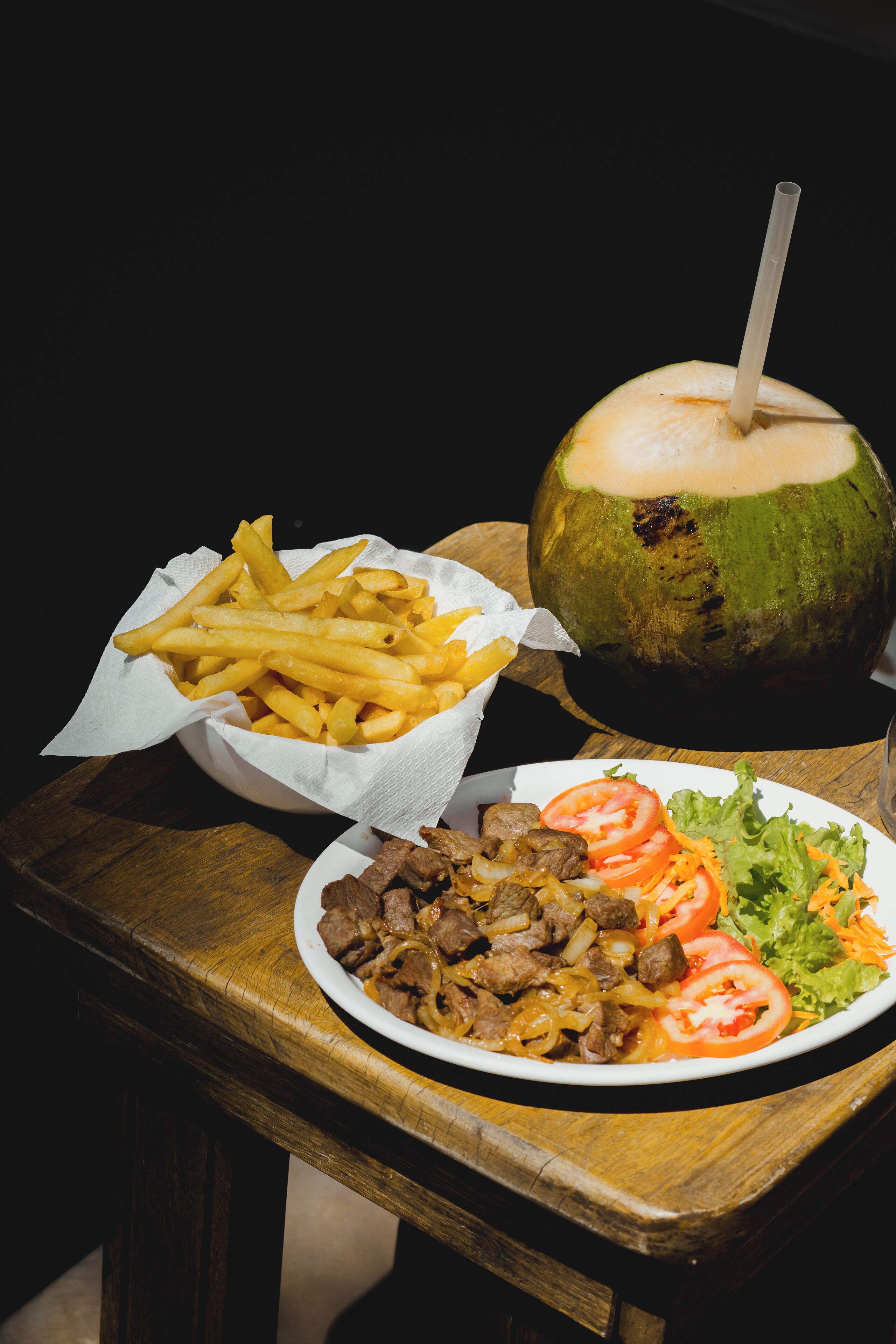 Una mesa de madera con un plato de comida, una cesta de papas fritas y un coco con bebida.