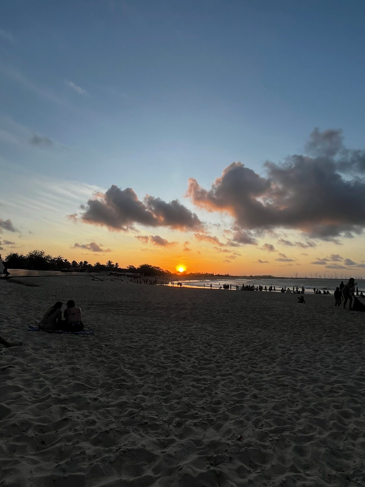 Uma praia ao pôr do sol com pessoas e um píer ao fundo.