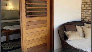 Une pièce avec une porte coulissante en bois, un lit avec une literie blanche, une chaise en osier avec des coussins et un mur en briques.