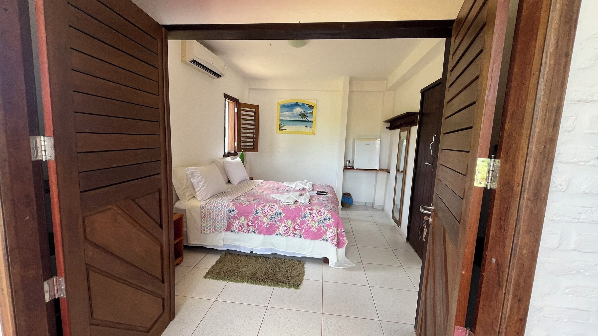 A bedroom with a bed, a small bedside table, a nightstand, a window with blinds, a wall-mounted air conditioner, a painting, and a door with a metal handle.