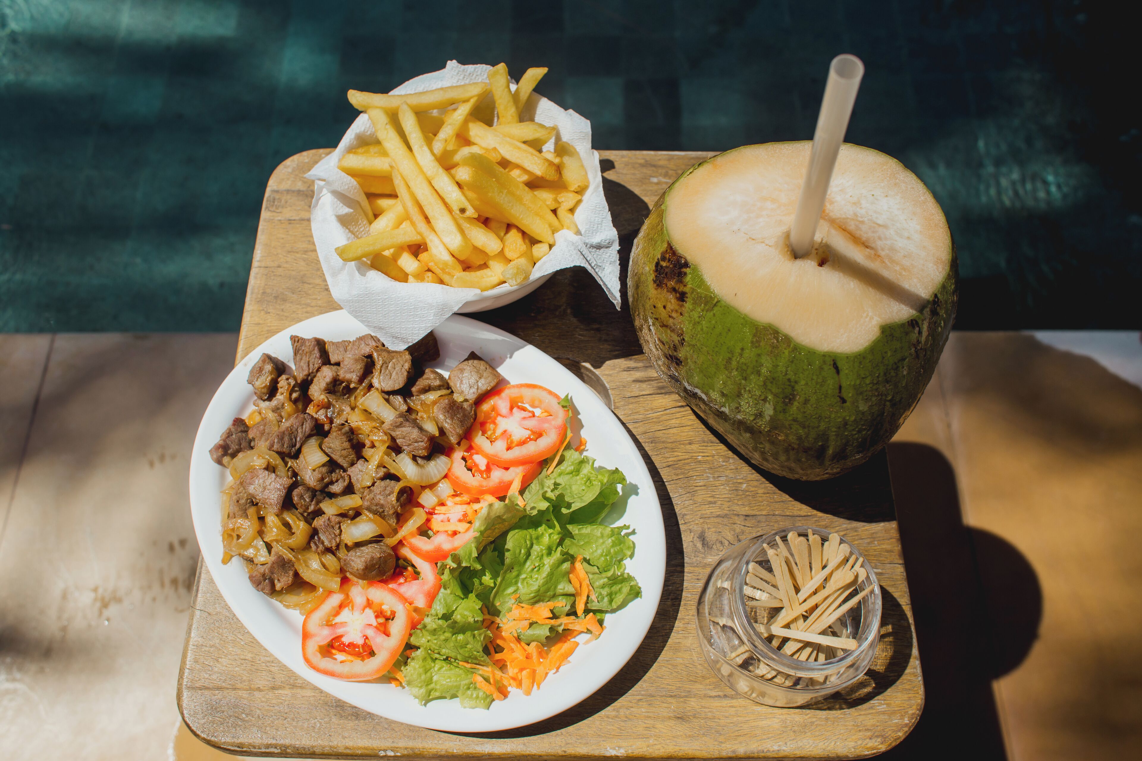A meal consisting of a plate of meat with onions and tomatoes, a bowl of French fries, a coconut drink, and a small bowl of toothpicks on a wooden table.