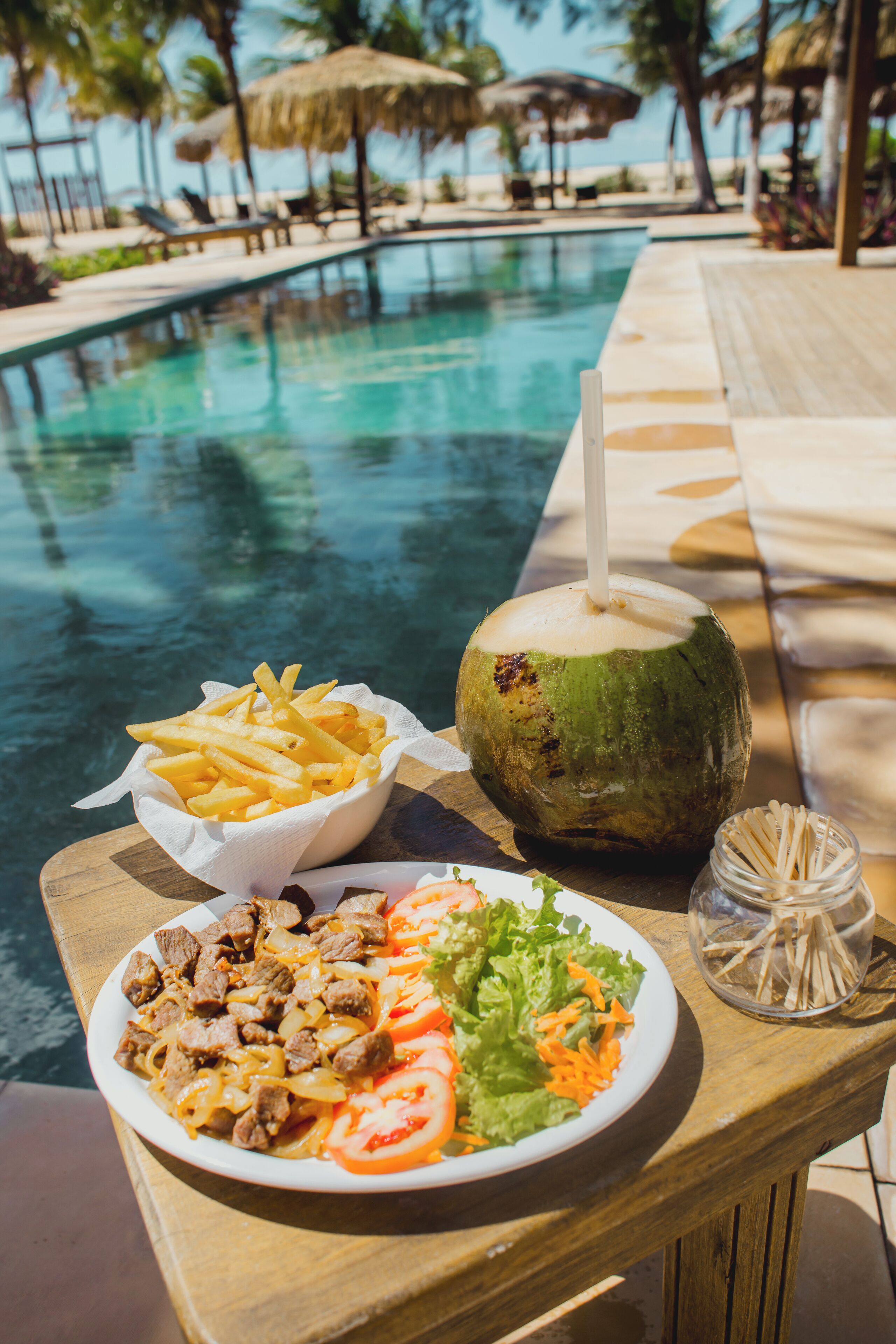 A dining table with a plate of food, a coconut drink, and a glass of straws by a poolside.