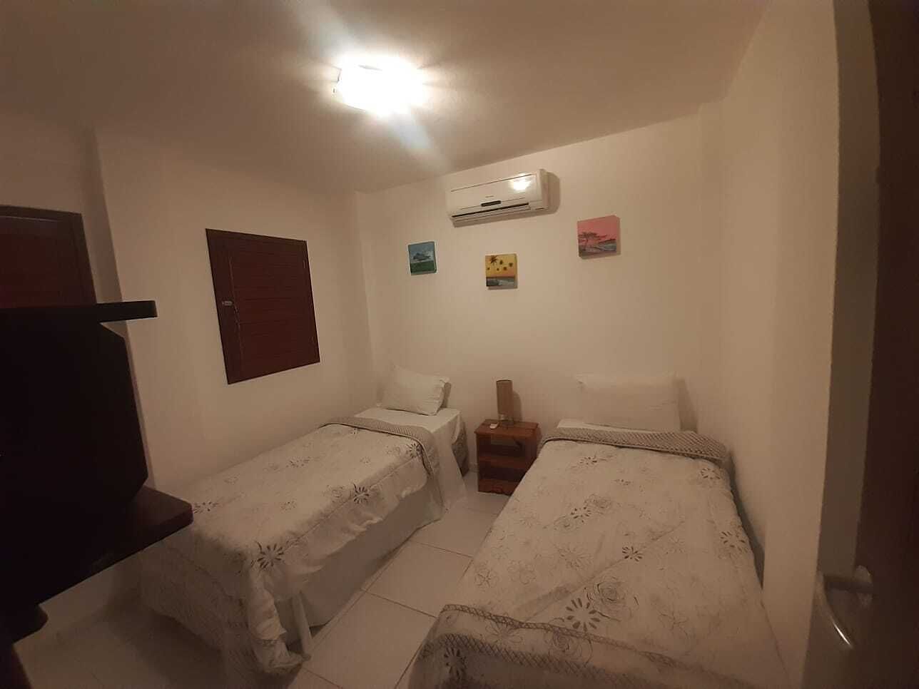 A small, dimly lit room with two single beds, a wooden nightstand, and a wall-mounted air conditioner.