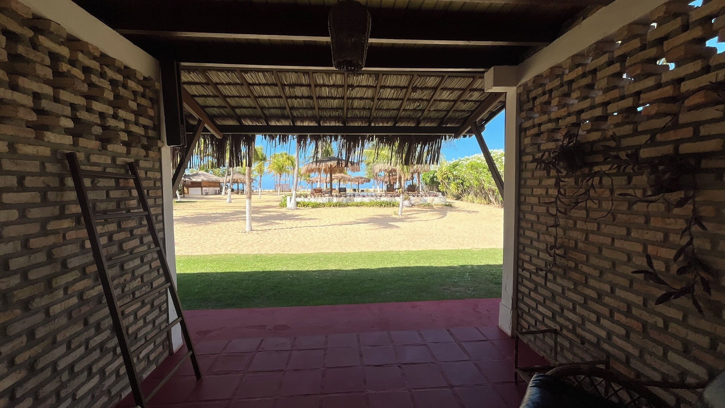 A covered outdoor area with a brick wall, a thatched roof, and a view of a beach with palm trees and a thatched-roof structure.