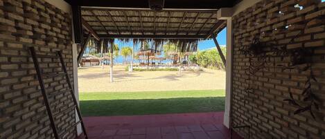 A covered outdoor area with a brick wall, a thatched roof, and a view of a beach with palm trees and a thatched-roof structure.
