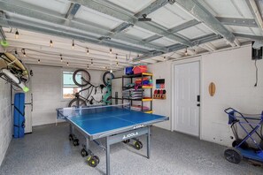 Une table de ping-pong dans une pièce dont le plafond est en métal, avec des vélos et des étagères remplies d’articles de sport.