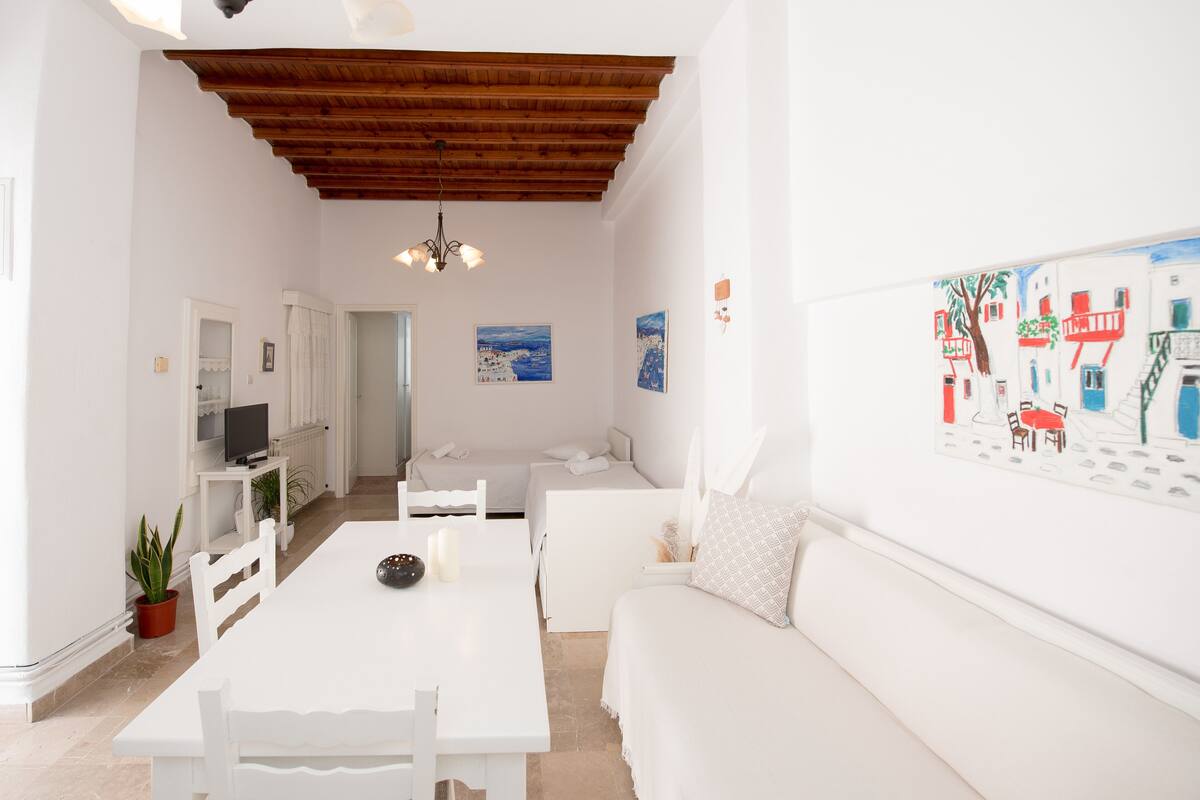 Un interior luminoso y blanco con techo de madera, una mesa de comedor, un sofá y un televisor sobre un soporte blanco.