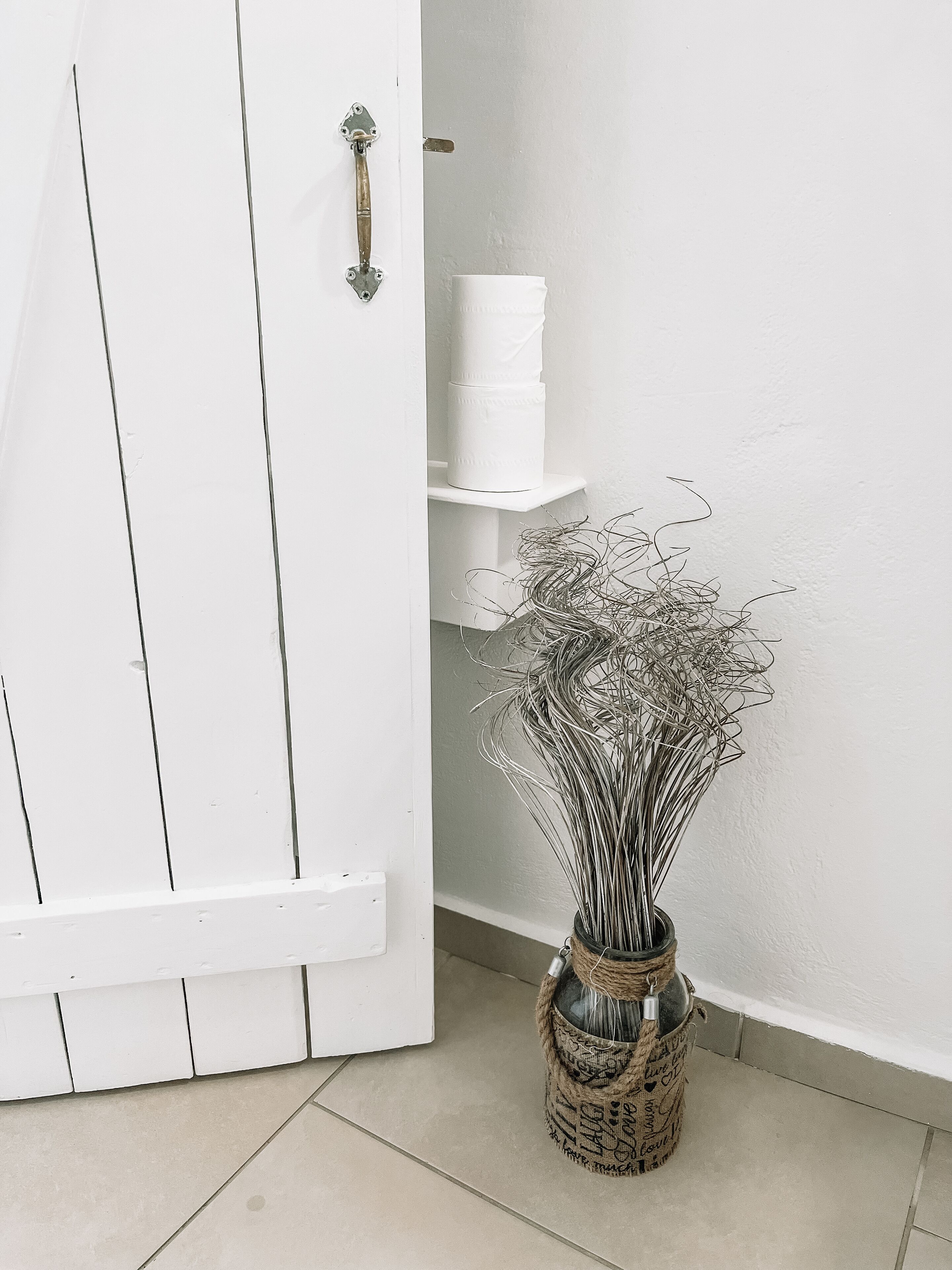 Uma porta branca com puxador de metal, uma prateleira branca com recipientes brancos empilhados e uma cesta de vime com uma planta em arame.