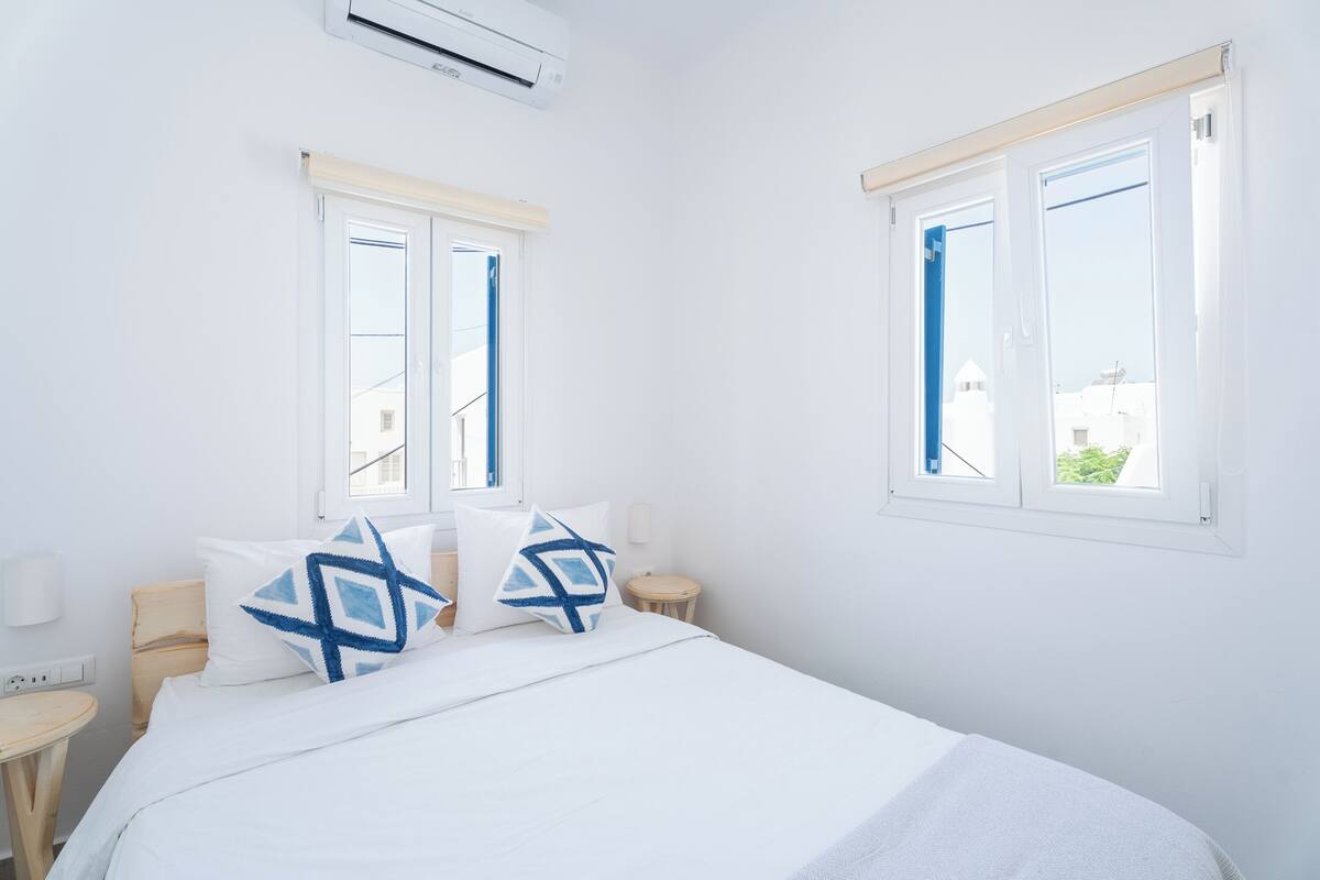 Una habitación luminosa y blanca con una cama, dos ventanas y una mesita de noche pequeña.