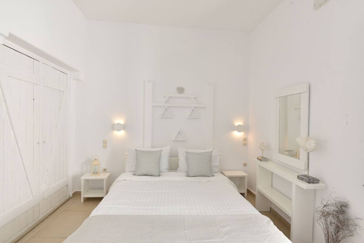 Un dormitorio blanco con una cama grande, mesitas de noche, un espejo y un armario.