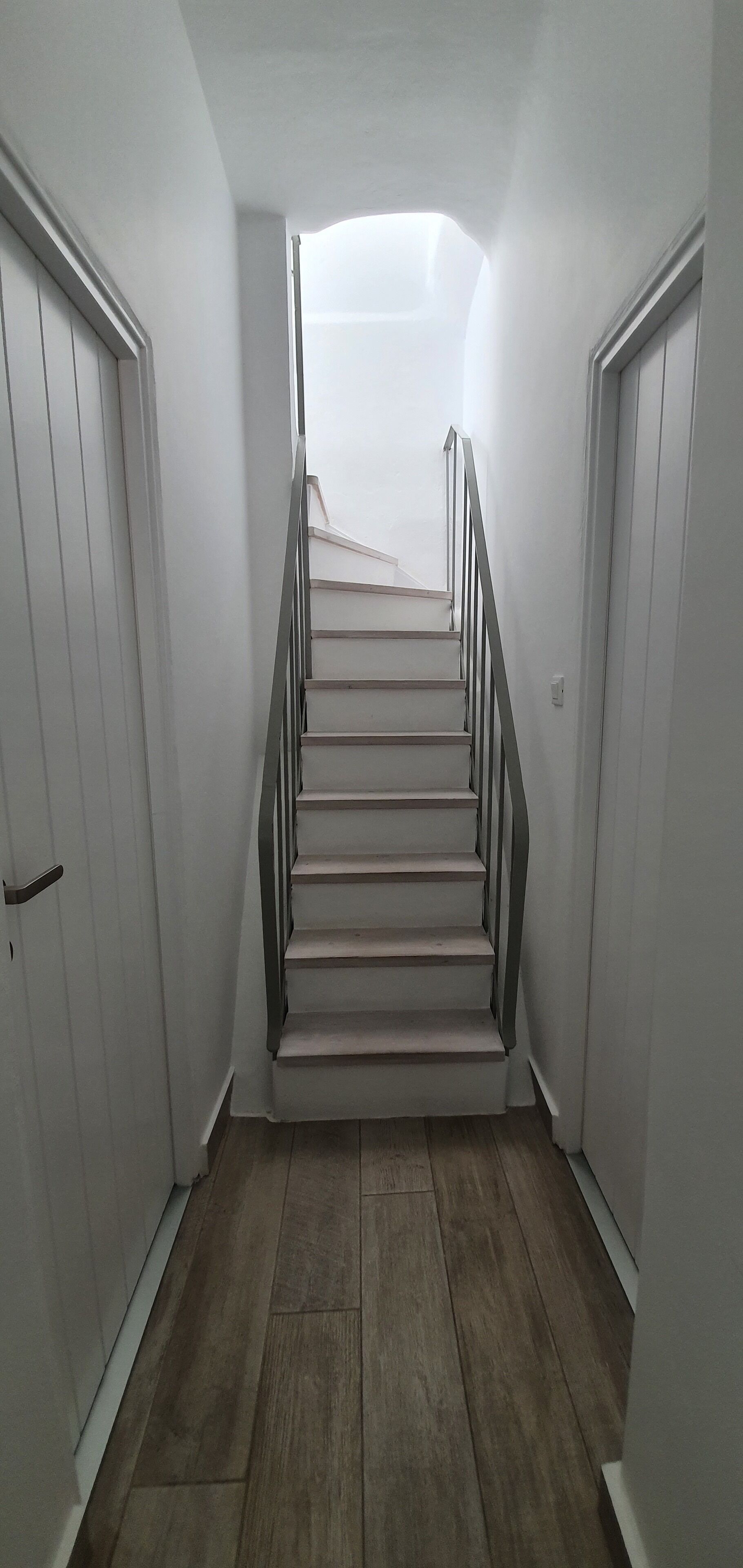 Un escalier doté d’une rampe, bordé de murs blancs et recouvert d’un plancher en bois, menant à un niveau supérieur.