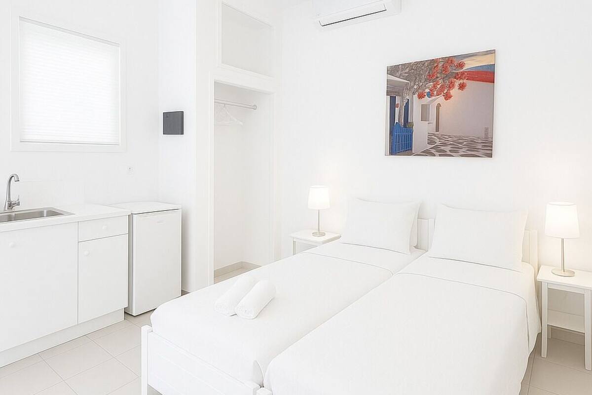 Una habitación blanca con una cama, una mesita, una mesita de noche, un ventanal y un cuadro en la pared.