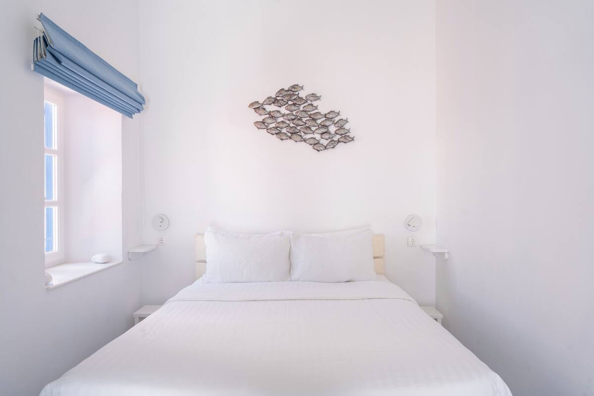 Una habitación blanca con una cama, una ventana con cortinas azules y una lámpara fijada a la pared.