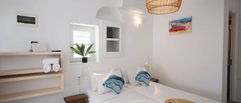 A bedroom with a bed, a bedside table, a shelf with towels, a window, a plant, and a painting.