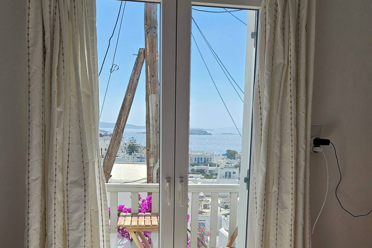 Un dormitorio con cama, un balcón con mobiliario de madera y vistas a edificios y agua.