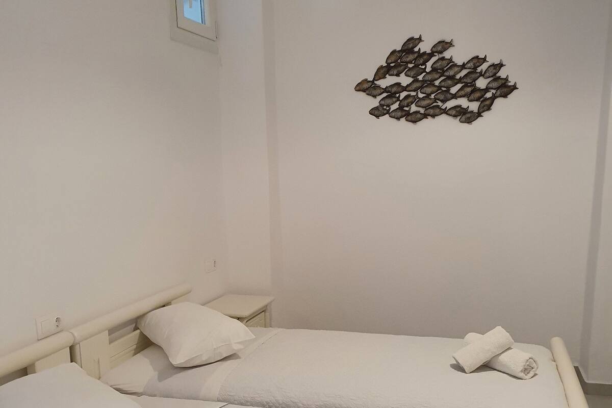 Una habitación con dos camas, ropa de cama blanca, un ventanal pequeño y un adorno de peces en la pared.