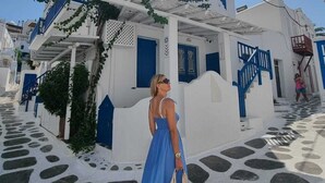 A white building with blue accents, a stone-paved street, and a person in a blue dress carrying a bag. - Eleanna's Mykonos (Mykonos)