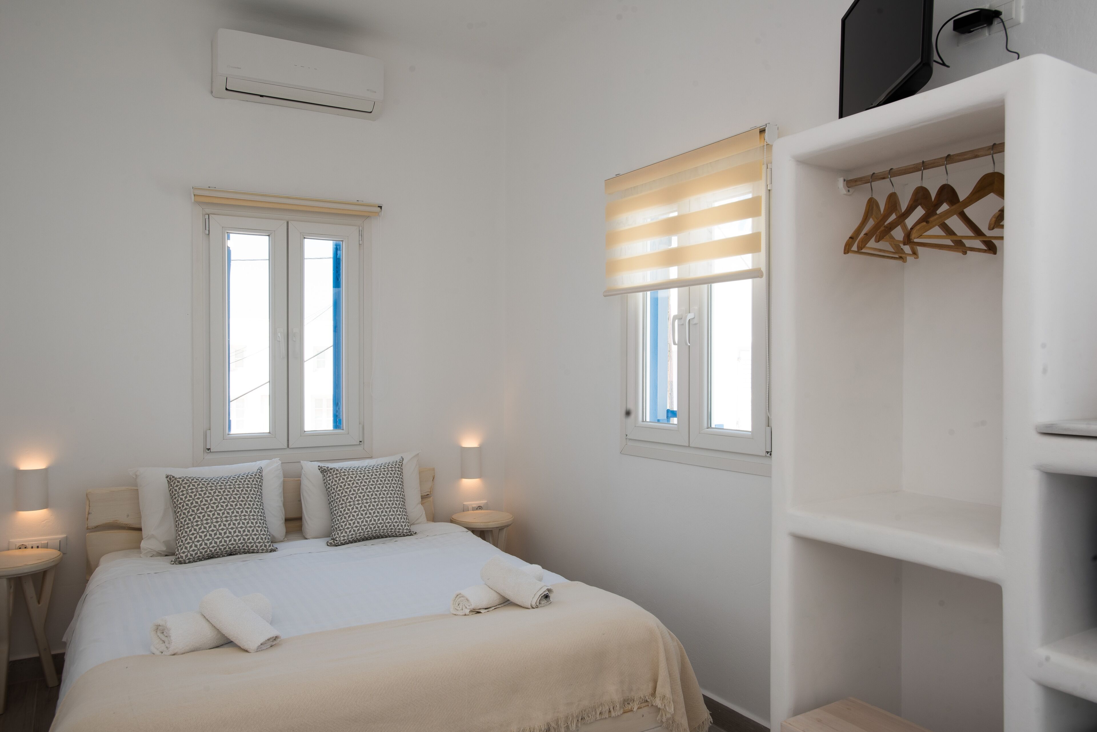 A bedroom with a bed, two windows, a bedside table, a lamp, and a wardrobe.