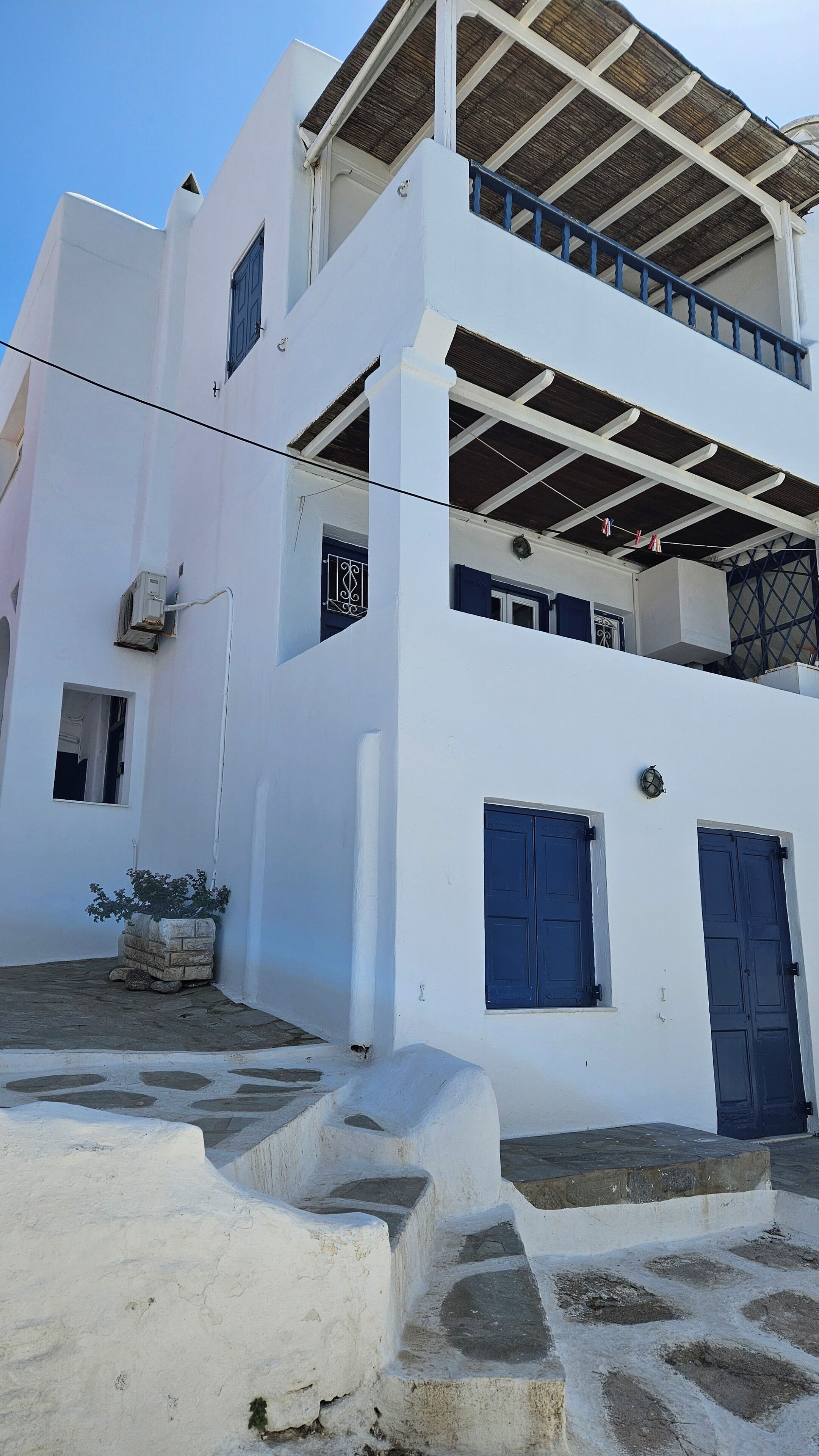 Un bâtiment blanc avec des portes et des fenêtres bleues, un balcon doté d’un plafond en bois et un escalier en pierre menant à l’entrée.