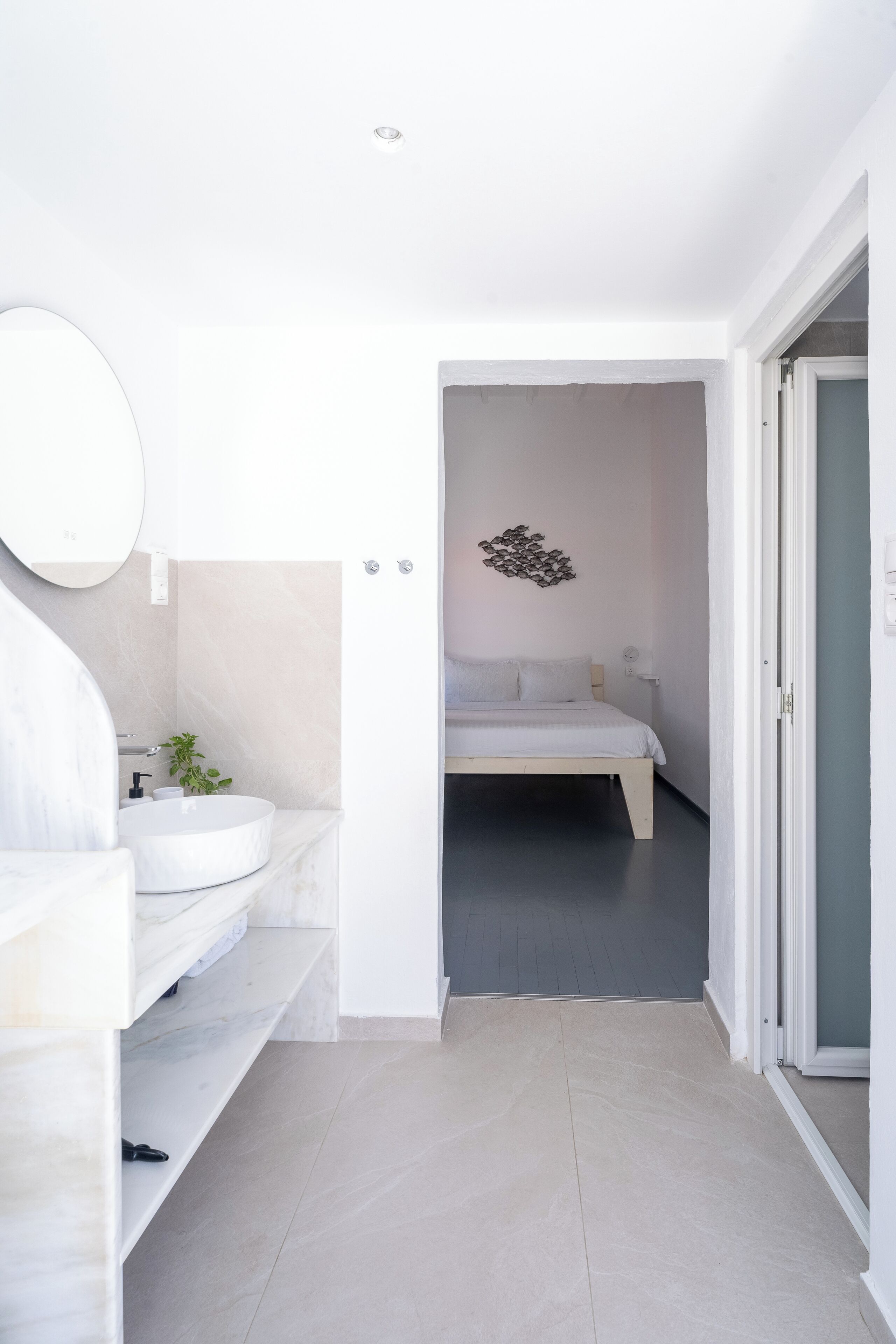 Un baño moderno con lavamanos y espejo blancos, y una cama en la habitación contigua.