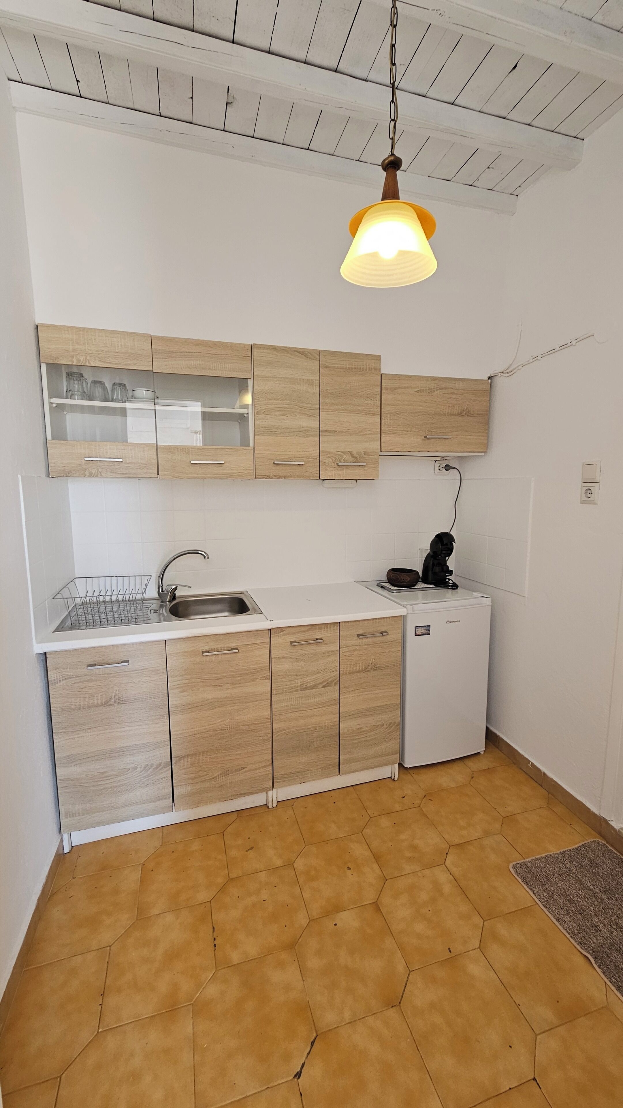 Kompakti keittiö, jossa on puiset kaapit, valkoinen työtaso, pesuallas ja pieni jääkaappi.
